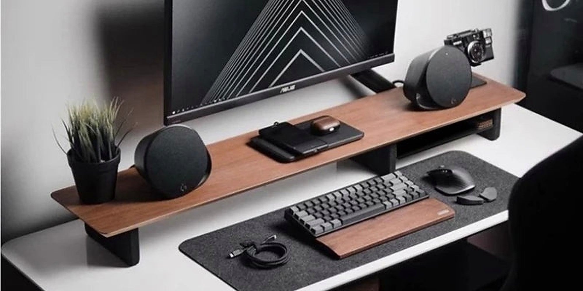 A walnut monitor shelf spanning a white desk, with a monitor, speakers, mechanical keyboard and felt desk mat